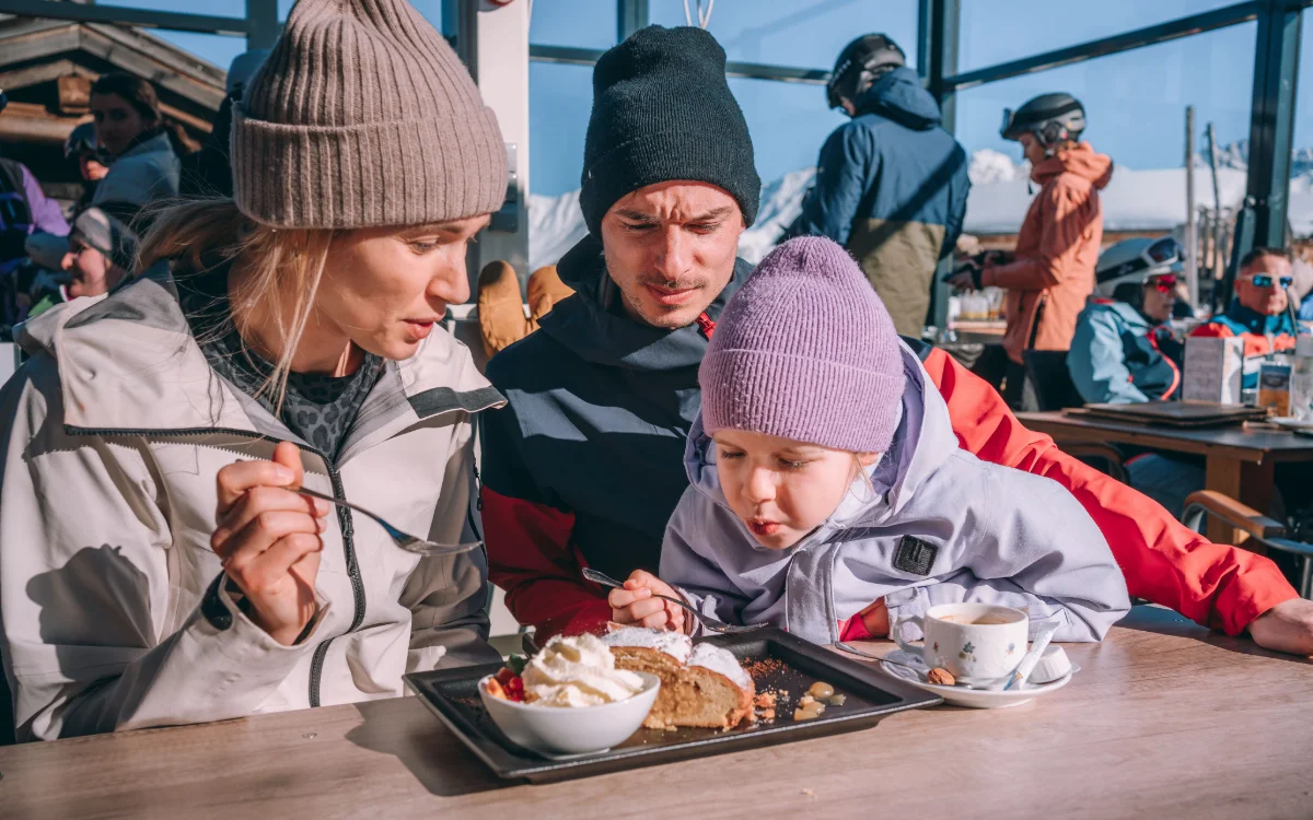 Eine Frau, ein Mann und ein Kind sitzen an einem Tisch auf einer Terrasse und essen Kuchen und andere Süßspeisen.