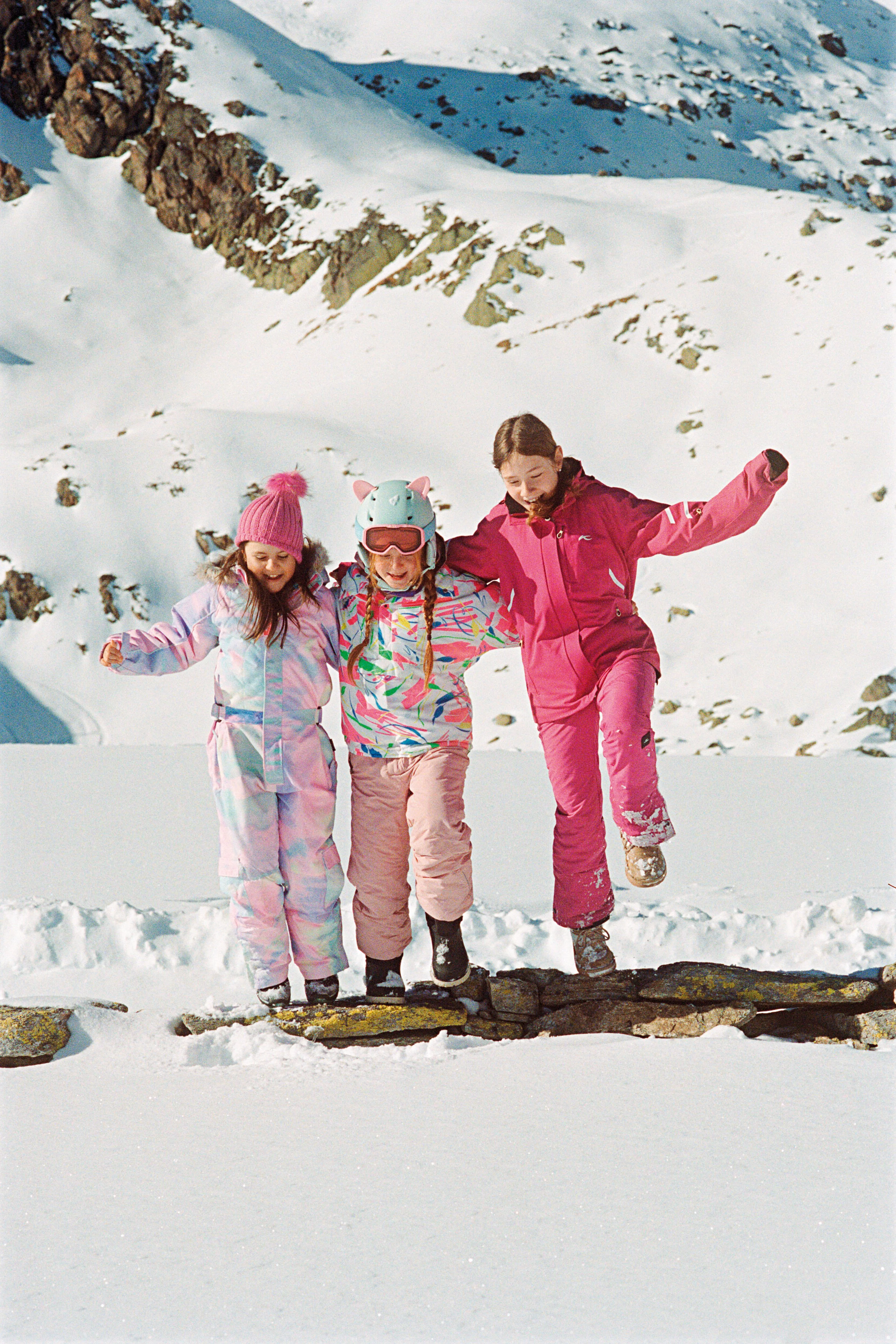 Drei Mädchen in bunter Skikleidung stehen nebeneinander im Schnee und halten die Arme ausgebreitet. Hinter ihnen liegt ein schneebedeckter Berghang.