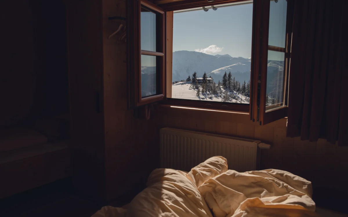 Ein zerwühltes Bett steht in einem dunklen Zimmer vor einem geöffneten Fenster mit Blick auf verschneite Berge und Nadelbäume.
