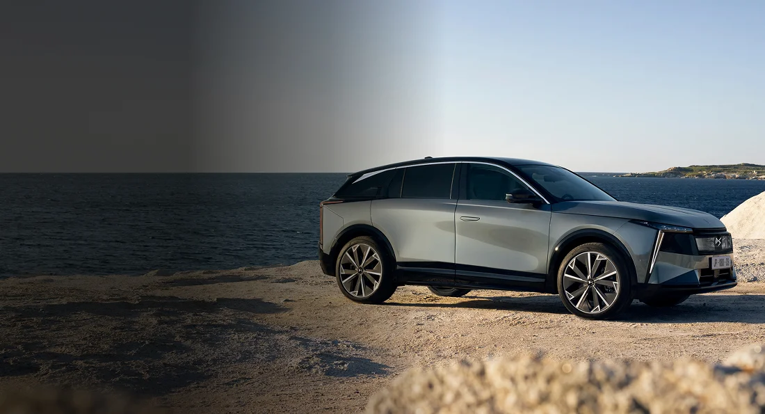 Grauer SUV an einer felsigen Küste vor dunklem Meer und blauem Himmel.