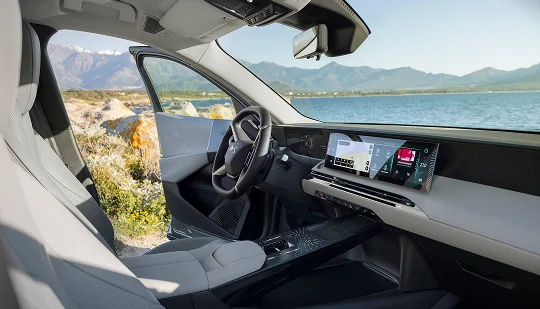 Innenraum eines Autos mit Lenkrad, digitalen Bildschirmen und geöffnetem Seitenfenster; dahinter sind Berge und ein See zu sehen.