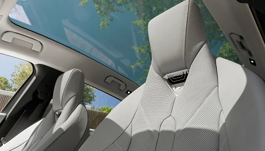 Zwei helle Vordersitze in einem Auto unter einem großen Glasdach. Durch das Dach und die Fenster sind Bäume und blauer Himmel sichtbar.
