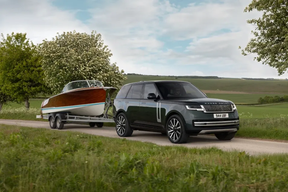 Ein dunkelgrüner SUV zieht auf einer Landstraße ein Holzboot mit weißem Rumpf auf einem Anhänger. Im Hintergrund stehen blühende Bäume und eine grüne Hügellandschaft.