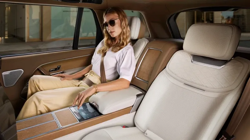 Eine Frau mit Sonnenbrille und weißem T-Shirt sitzt auf dem beigen Rücksitz eines Autos und bedient ein Display in der Mittelkonsole. Die Sitze und Verkleidungen sind in Beige und Braun gehalten.
