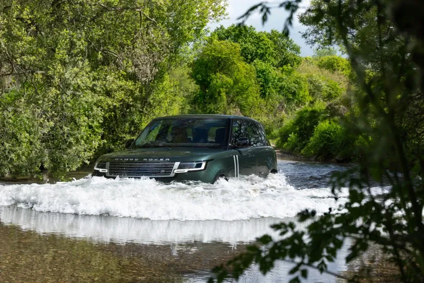 Ein dunkelgrüner Geländewagen fährt durch einen flachen Fluss, vor dem Fahrzeug spritzt weißes Wasser auf. Am Ufer stehen grüne Bäume und Büsche.